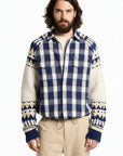 A male model wearing a blue and cream plaid button-up shirt layered over a cream knit sweater, paired with beige drawstring pants, against a plain white background.