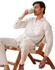 Man sitting on a chair wearing a white embroidered shirt and beige pants, drinking from a cup.