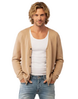 A man modeling a beige knit cardigan over a white tank top and light blue distressed jeans, standing against a light gray studio background.