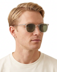Blonde male model wearing Garrett Leight Bentley sunglasses with clear Bio Glass frames and Bio Green lenses on a white background