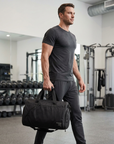 Model carrying the Mack Weldon ATLAS duffle in Asphalt beside dumbbell racks and gym equipment”
