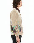 A man shown in profile wearing a light beige denim jacket with colorful abstract graffiti-style paint splatters on the lower front and sleeves, paired with white pants against a plain white background.