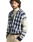 A young man wearing a blue and white plaid button-up shirt layered over a cream knit sweater with blue and cream Fair Isle patterned sleeves and cuffs, paired with beige pants and a brown belt, against a solid white background.