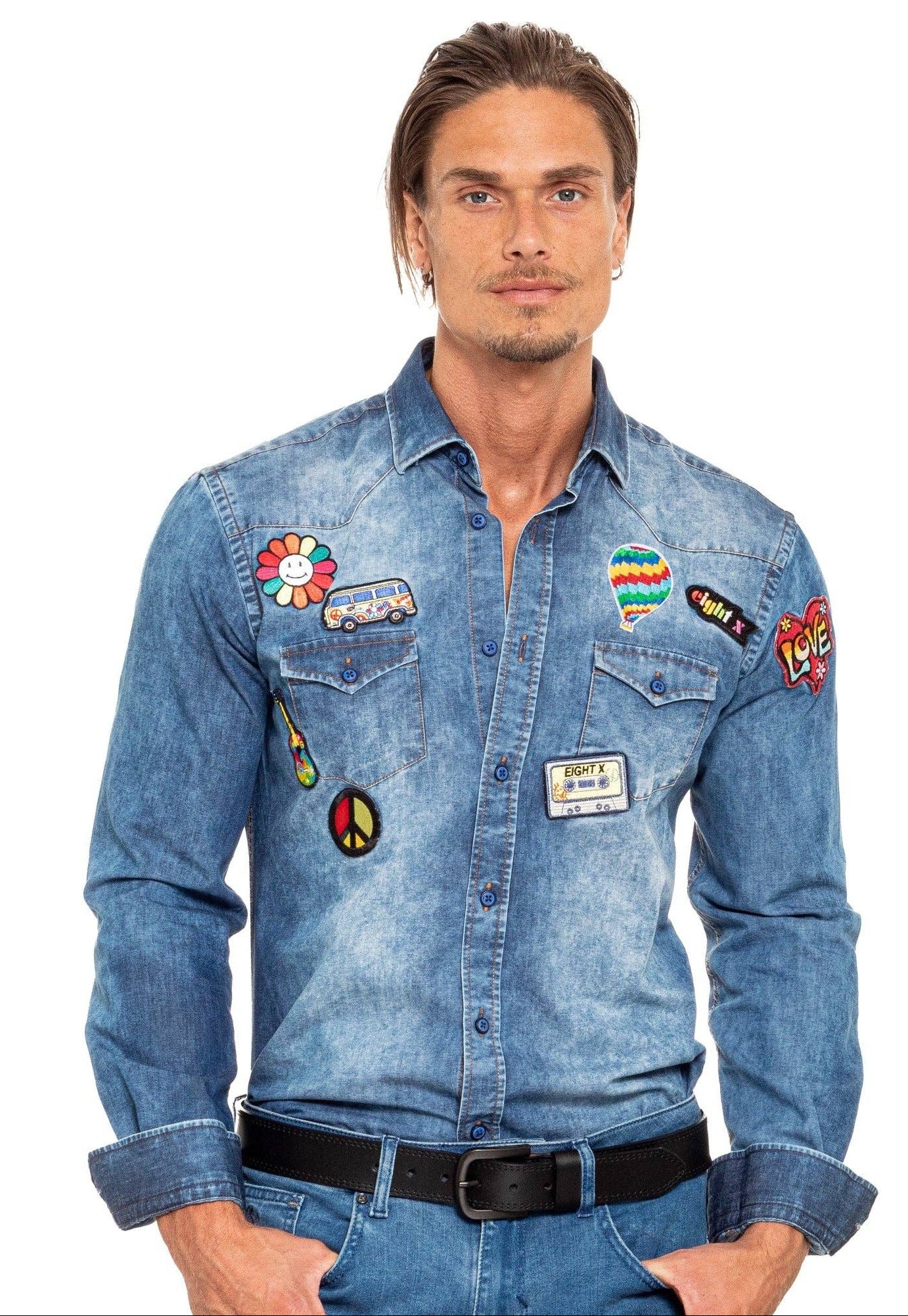 A man wearing a light blue denim shirt jacket decorated with multiple colorful embroidered patches, paired with blue jeans and a black belt, against a plain white background.