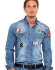 A man wearing a light blue denim shirt jacket decorated with multiple colorful embroidered patches, paired with blue jeans and a black belt, against a plain white background.