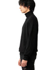 A model with black wavy hair is shown in profile wearing a black turtleneck sweater and black distressed jeans, against a plain white studio background.