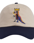 A structured baseball cap with a beige crown and a navy blue curved brim, featuring an embroidered cartoon dinosaur wearing a gold crown on the front.
