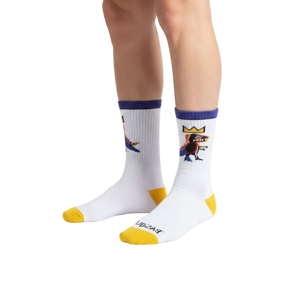 A pair of white crew socks with yellow toes and heels, purple cuffs, and a colorful cartoon graphic on the upper right sock. The brand name 'BROOKS' is printed in black on the sole of the front sock.